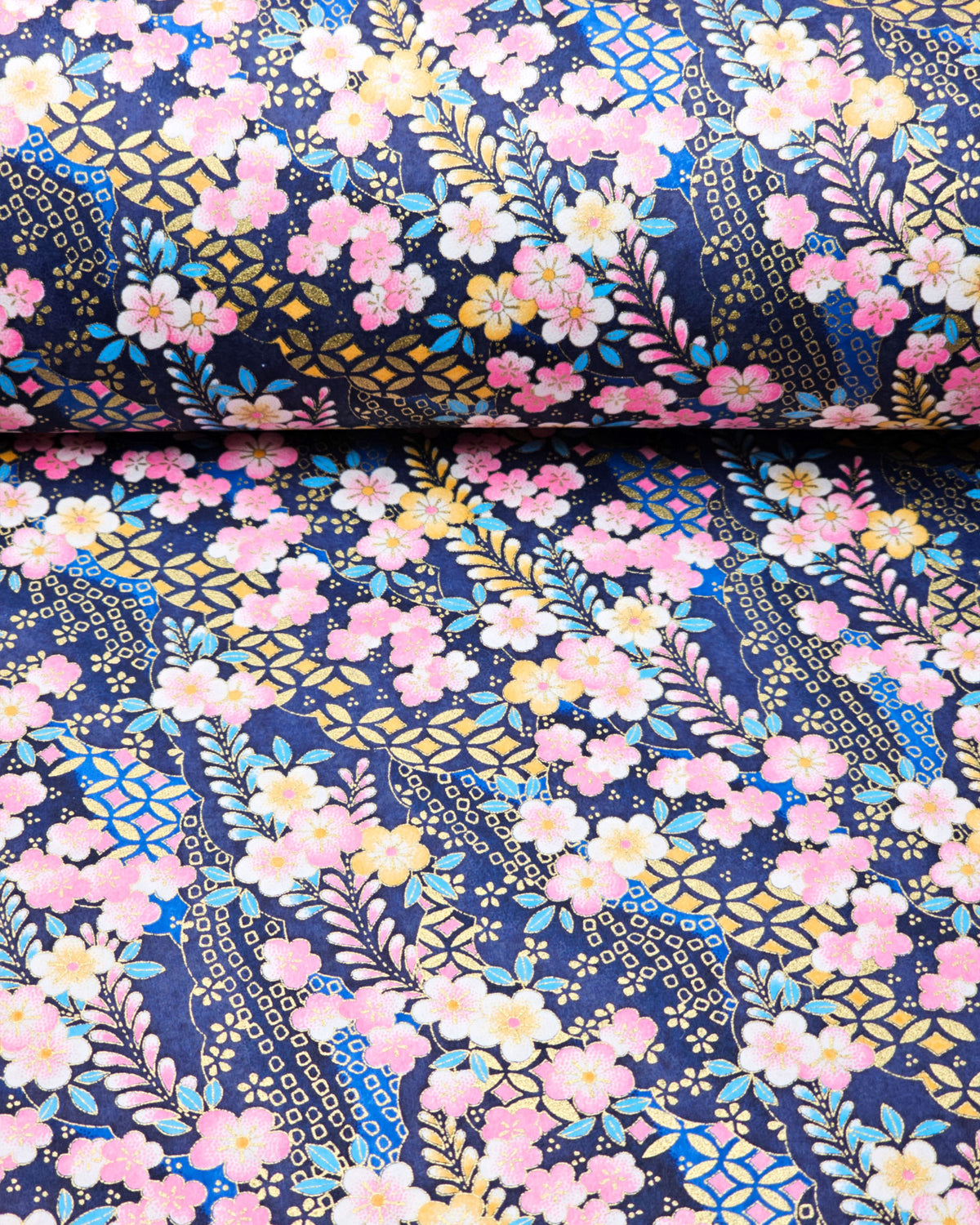 Japanese chiyogami paper featuring ypink and yellow plum blossoms and blue, pink, and yellow wisteria on a dark blue background with metallic gold details, shown rolled to highlight the design.