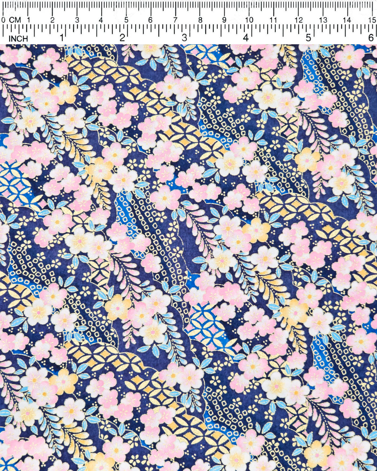 Japanese chiyogami paper featuring pink and yellow plum blossoms and blue, pink, and yellow wisteria on a dark blue background with metallic gold details, shown next to a ruler for size reference.