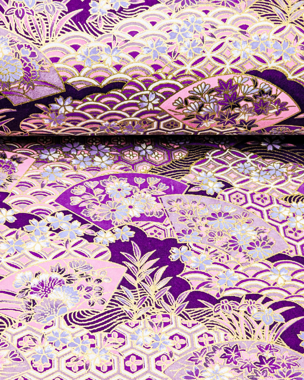 Japanese chiyogami paper featuring purple and pink floral hand fans and various traditional Japanese geometric motifs (shippo, kikko, seigaiha) on a dark purple background with metallic gold accents. Small purple and white cherry blossom are scattered throughout the design.