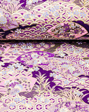Japanese chiyogami paper featuring purple and pink floral hand fans and various traditional Japanese geometric motifs (shippo, kikko, seigaiha) on a dark purple background with metallic gold accents. Small purple and white cherry blossom are scattered throughout the design.