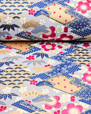 Japanese chiyogami paper featuring large pearlescent magenta pink and white plum blossoms, blue and gold bamboo leaves, and light blue and gold pine on a blue and gold background with metallic gold accents. The background is adorned with a various traditional Japanese geometric designs.