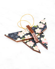 Small wooden origami butterfly ornament featuring pink and white cherry blossoms with green leaves on a black and gray asanoha background with metallic gold details.