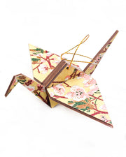 Large wooden origami crane ornament featuring pink and white plum blossoms with dark brown branches and green leaves on a beige background with metallic gold details.