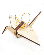 Small wooden origami crane ornament featuring small white and gold cherry blossoms on pearlescent cream background with metallic silver details.
