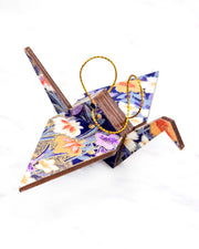 Small wooden origami crane ornament featuring orange, purple, and white irises on a dark blue background with metallic gold accents.