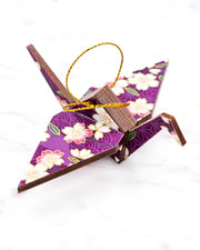 Small wooden origami crane ornament featuring pink and white cherry blossoms on a purple background with metallic gold accents.