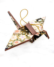 Small wooden origami crane ornament featuring pink, white, and purple cherry blossoms on a black background with metallic gold accents.