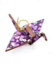 Small wooden origami crane ornament featuring purple and white plum blossoms with green branches on a black background with metallic gold accents.