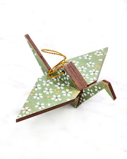 Small wooden origami crane ornament featuring small white and gold cherry blossoms on a light green background with metallic gold details.