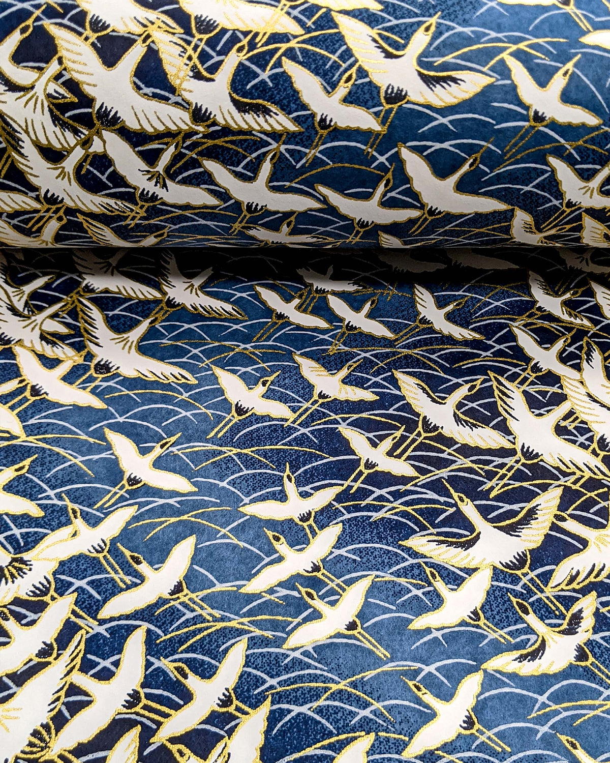 Japanese chiyogami paper featuring white flying cranes in a zigzag formation on a navy blue background with metallic gold details, shown rolled to highlight the design.