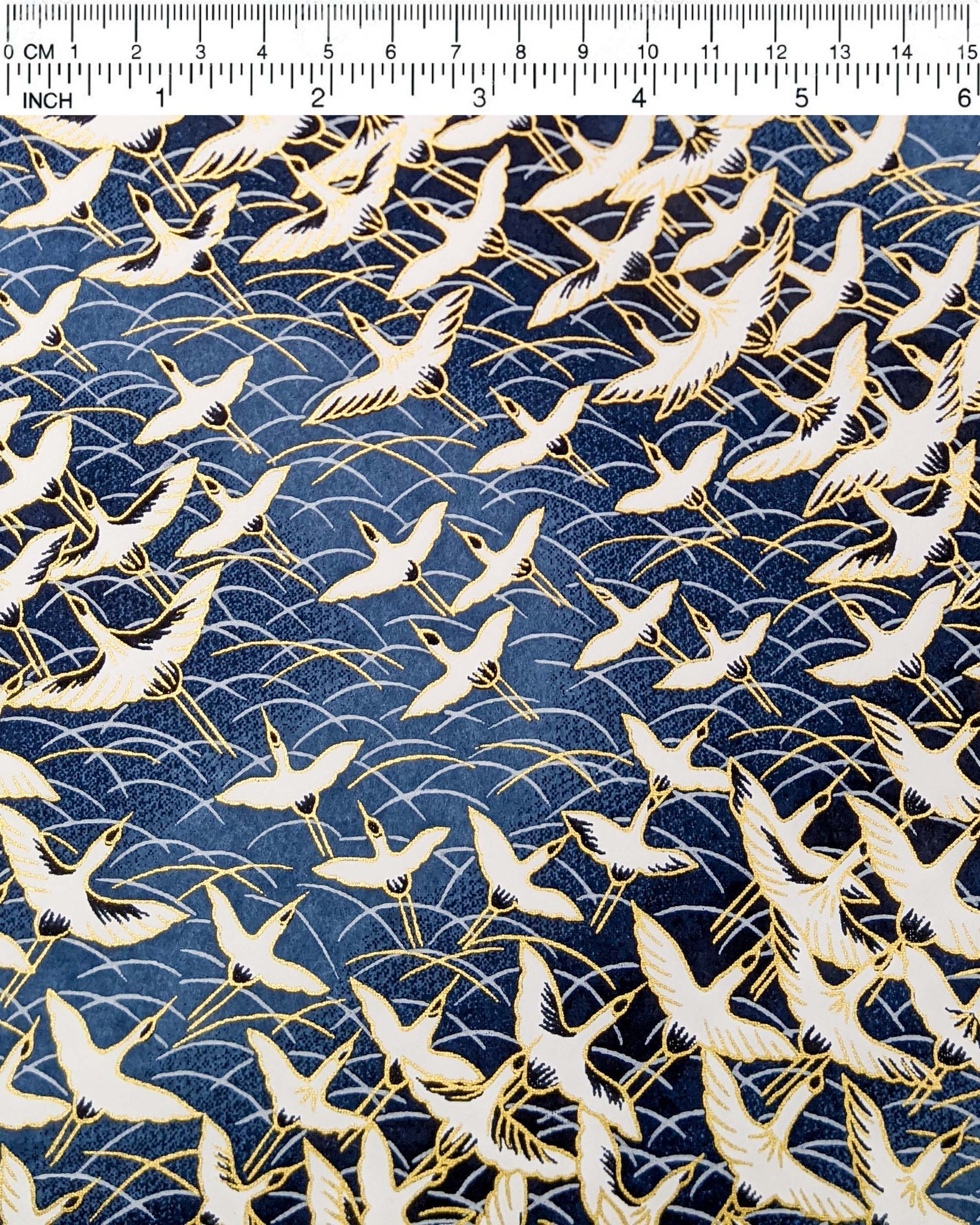 Japanese chiyogami paper featuring white flying cranes in a zigzag formation on a navy blue background with metallic gold details, shown next to a ruler for size reference.