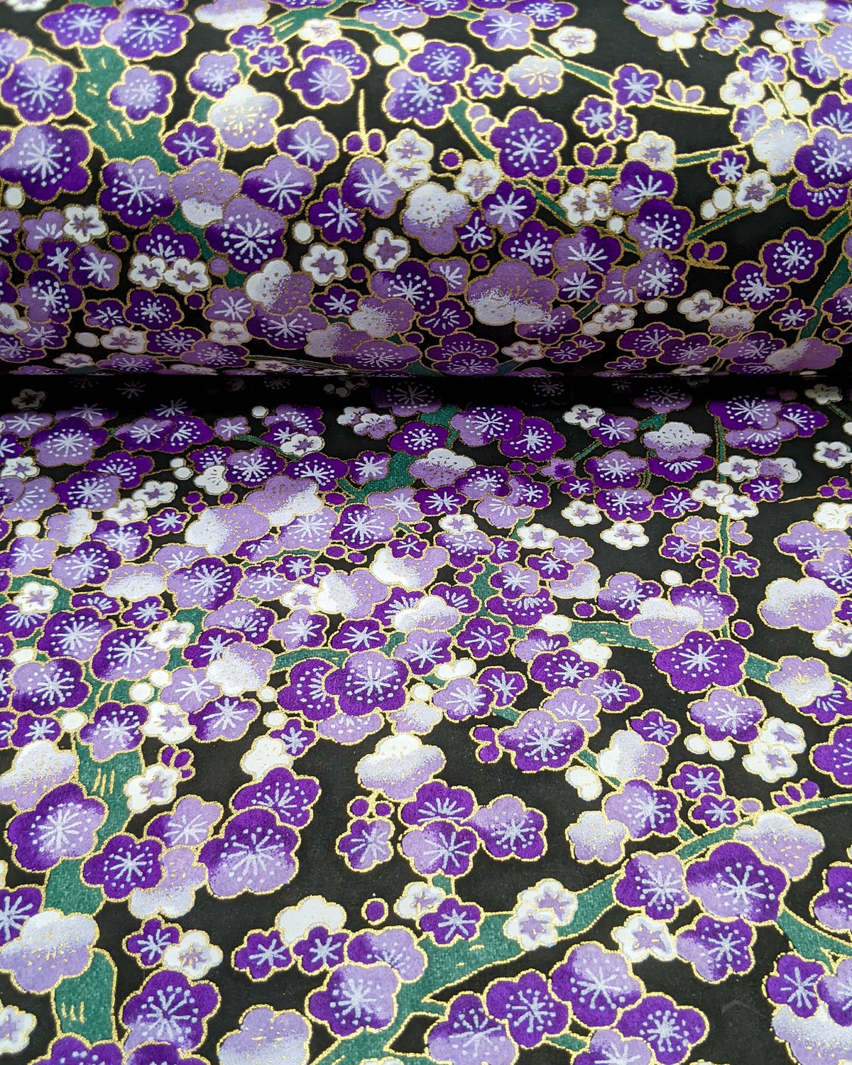 Japanese chiyogami paper featuring purple and white plum blossom trees with green branches on a black background with metallic gold details, shown rolled to highlight the design.