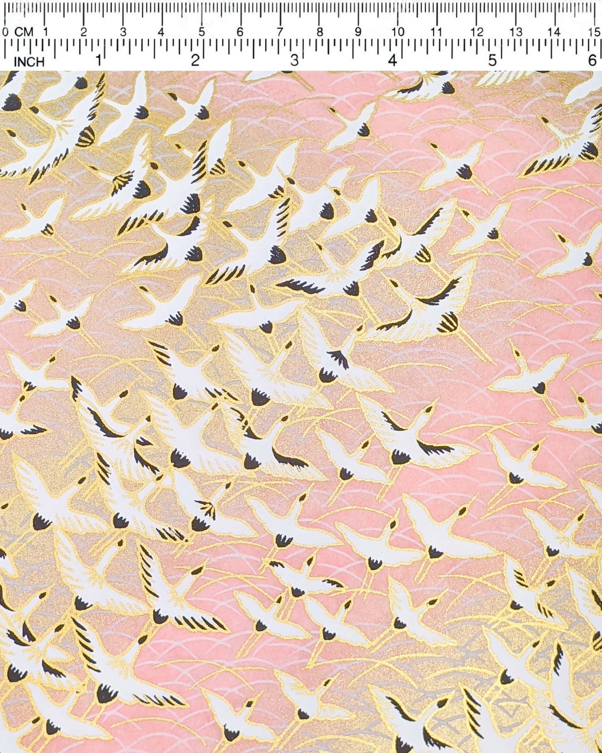 Japanese chiyogami paper featuring white flying cranes in a zigzag formation on a light pink background with metallic gold details.