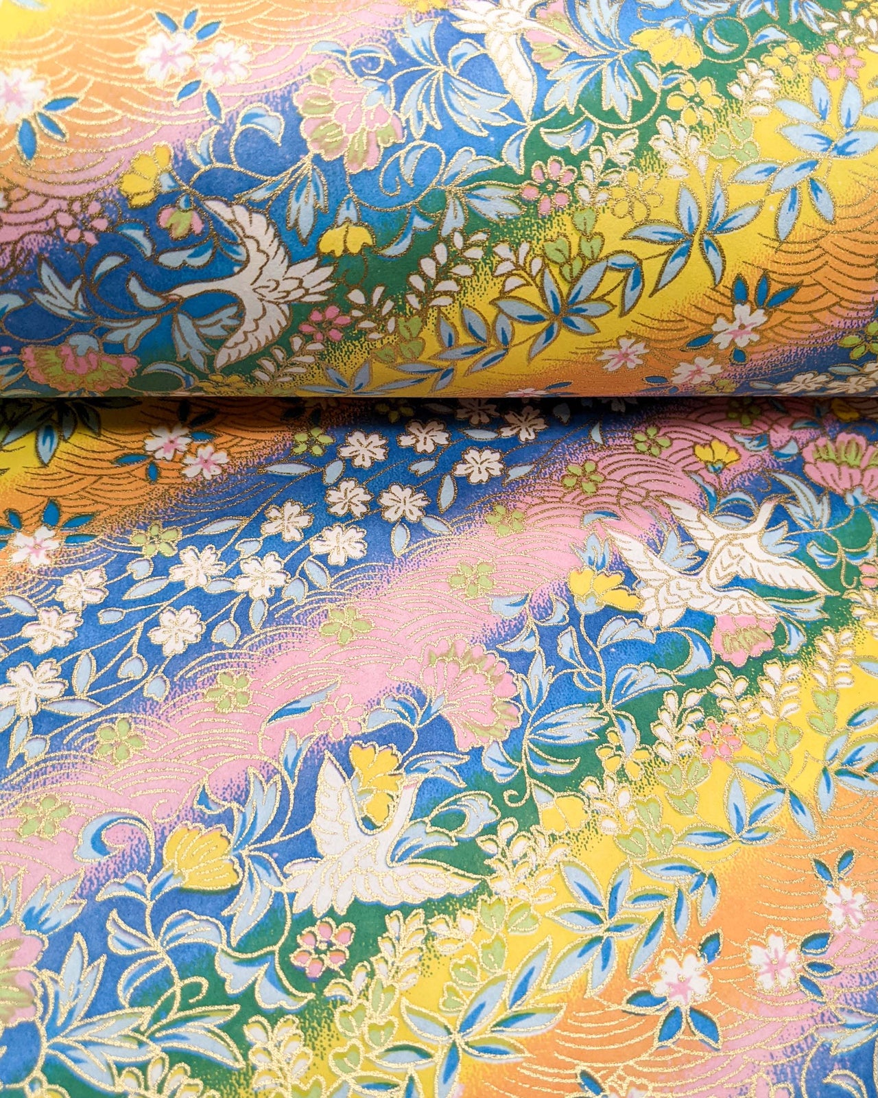 Japanese chiyogami paper featuring white cranes and a variety of flowers on a striped background in blue, pink, green, yellow, and orange with metallic gold details.