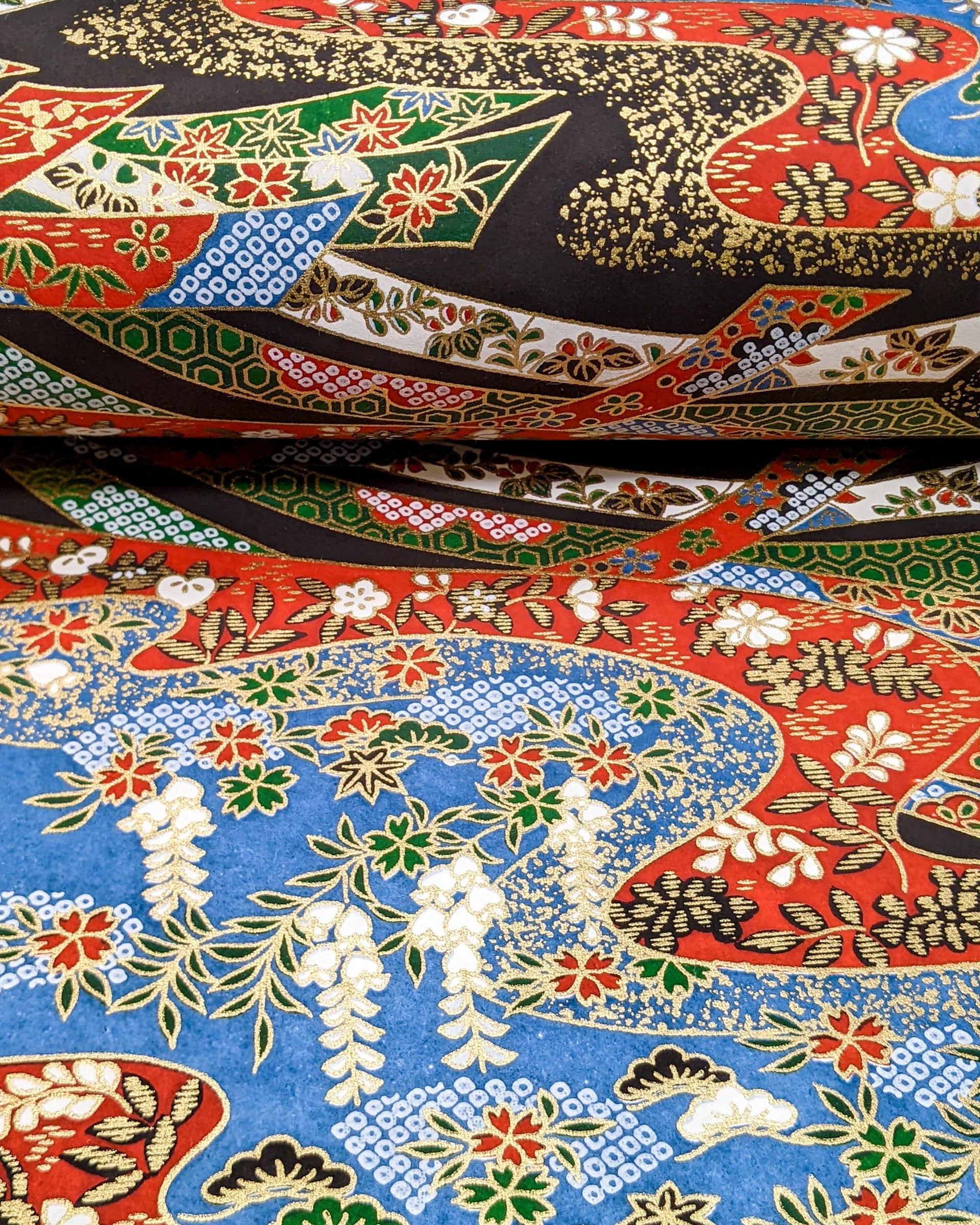 Japanese chiyogami paper featuring red, white, blue, and green ribbons decorated with traditional Japanese motifs on a black, blue, and red background with metallic gold details. The design includes scattered red and green cherry blossoms, green and black pines, red and black maples leaves, and white wisterias.