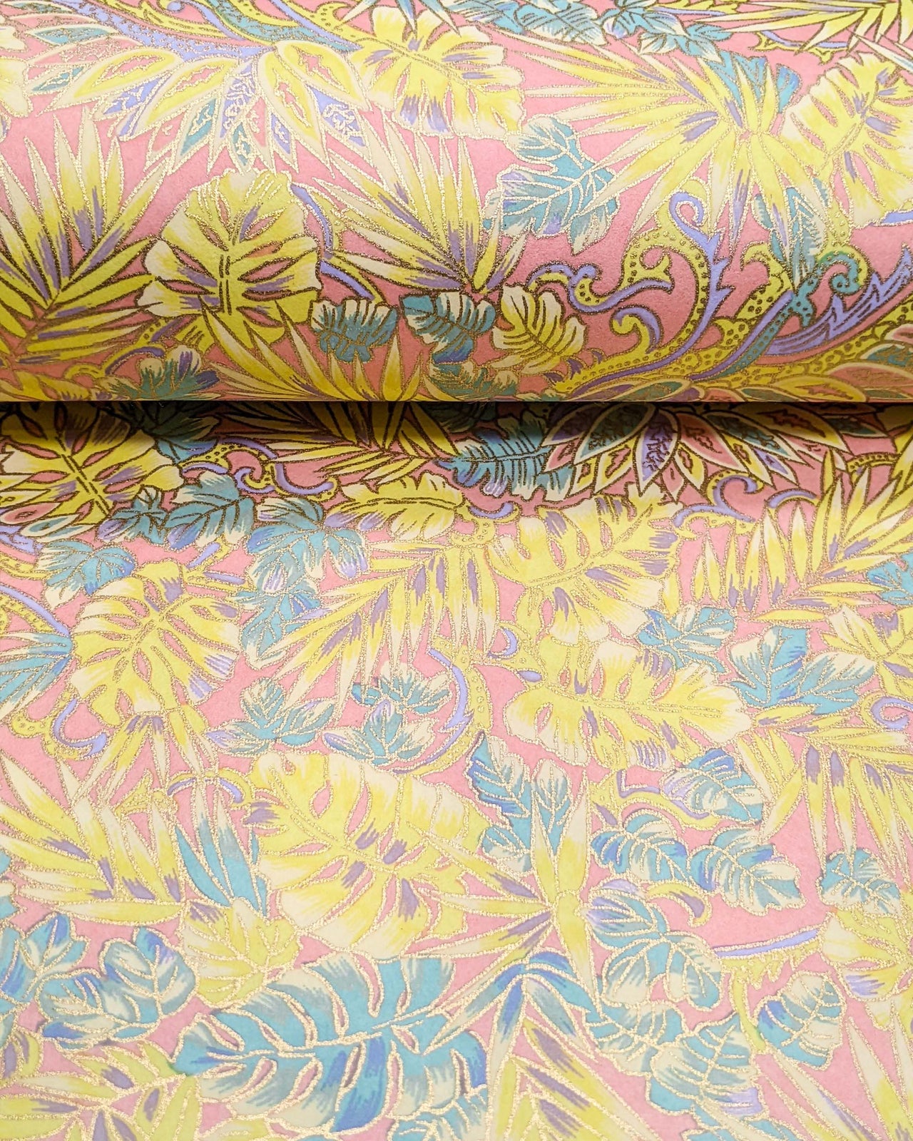 Japanese chiyogami paper featuring yellow, purple, and turquoise frond leaves on a light pink background with metallic gold accents, shown rolled to highlight the design.