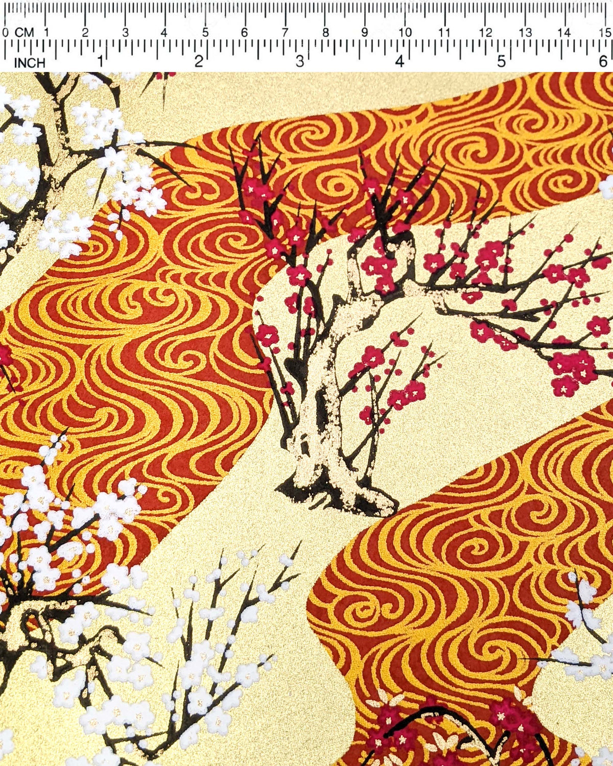 Japanese chiyogami paper featuring pink and white plum and cherry blossom trees set against a metallic gold and red water ripple pattern on a gold background, shown next to a ruler for size reference.
