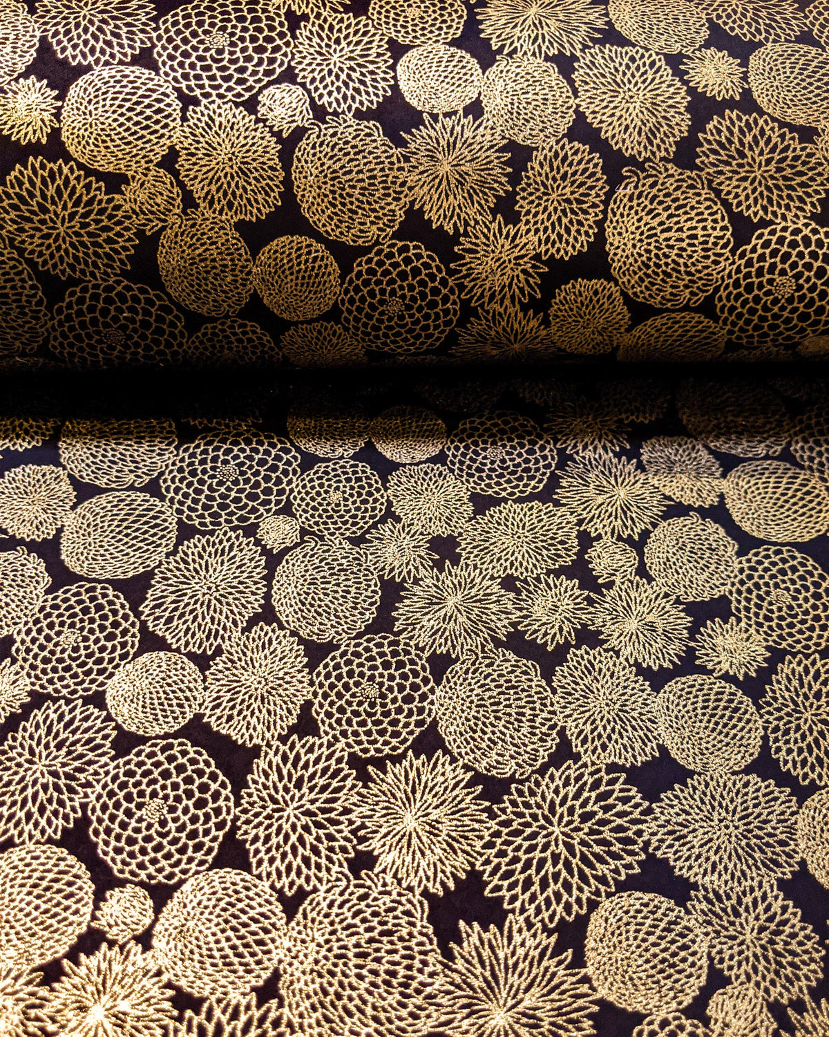 Japanese chiyogami paper featuring an assortment of small metallic gold chrysanthemums on a black background, shown rolled to highlight the design.