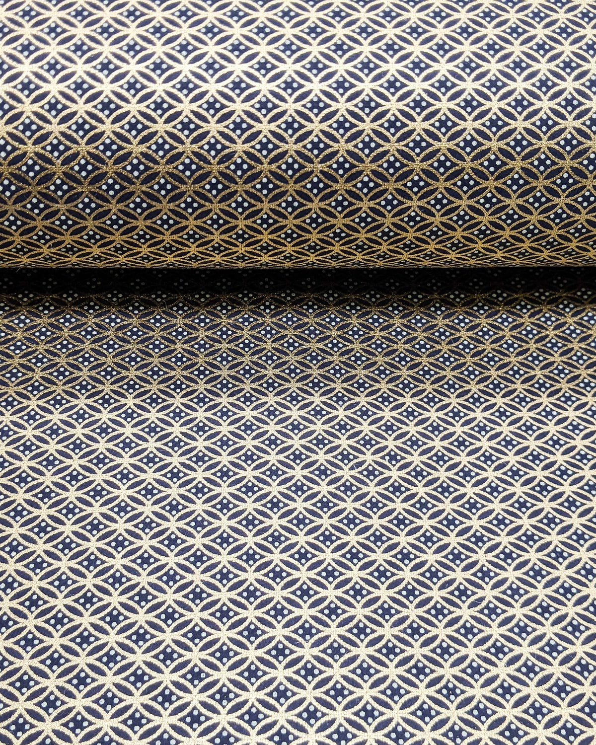 Japanese chiyogami paper featuring a traditional oval shippo pattern in metallic gold with four white dots in the center on a black background, shown rolled to highlight the design.