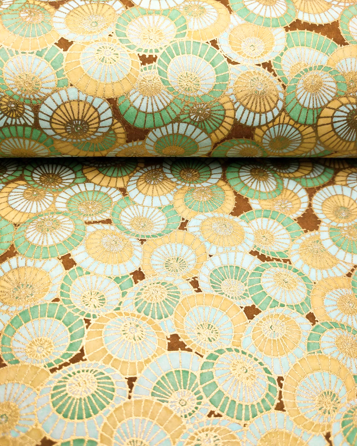 Japanese chiyogami paper featuring beige, turquoise, and light blue overlapping parasols on a brown background with metallic gold details, shown rolled to highlight the design.