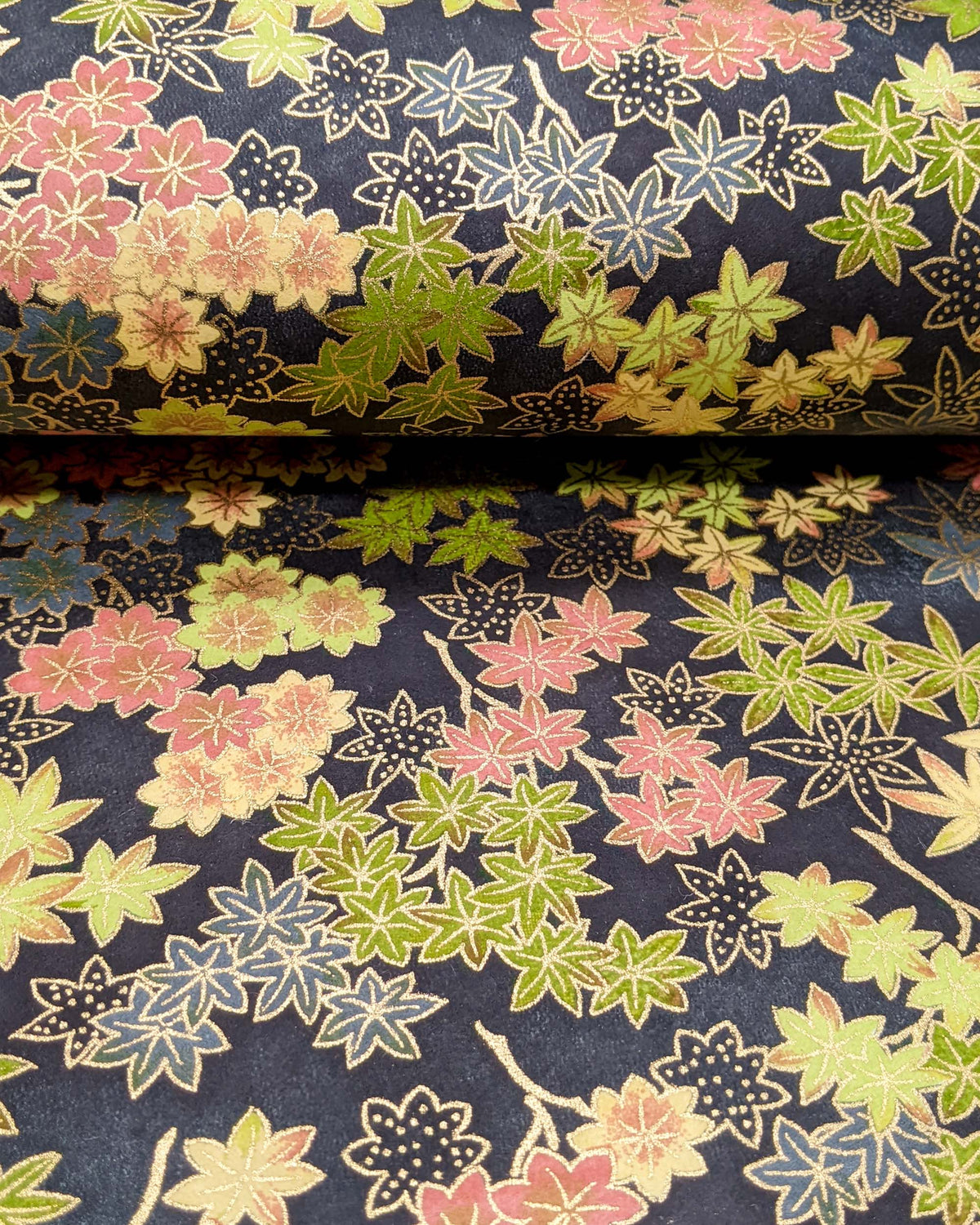 Japanese chiyogami paper featuring assorted green, pink, beige, and gray maple leaves with dark gray and metallic gold dotted maple leaves on a dark gray background, shown rolled to highlight the design.