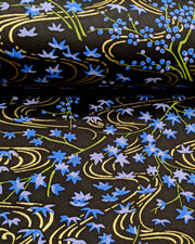 Japanese chiyogami paper featuring small blue maples leaves with green branches over flowing metallic gold streams on a black background, shown rolled to highlight the design.