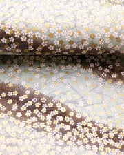 Japanese chiyogami paper featuring small white and metallic gold cherry blossoms on a gradient blue gray background, shown rolled to highlight the design.