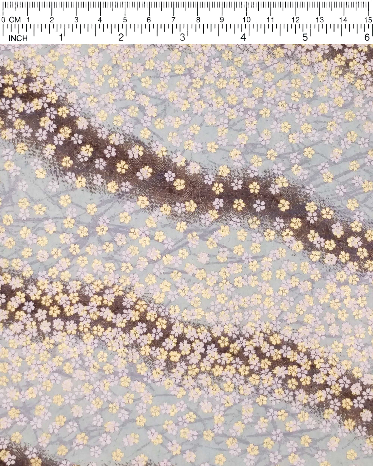 Japanese chiyogami paper featuring small white and metallic gold cherry blossoms on a gradient blue gray background, shown next to a ruler for size reference.