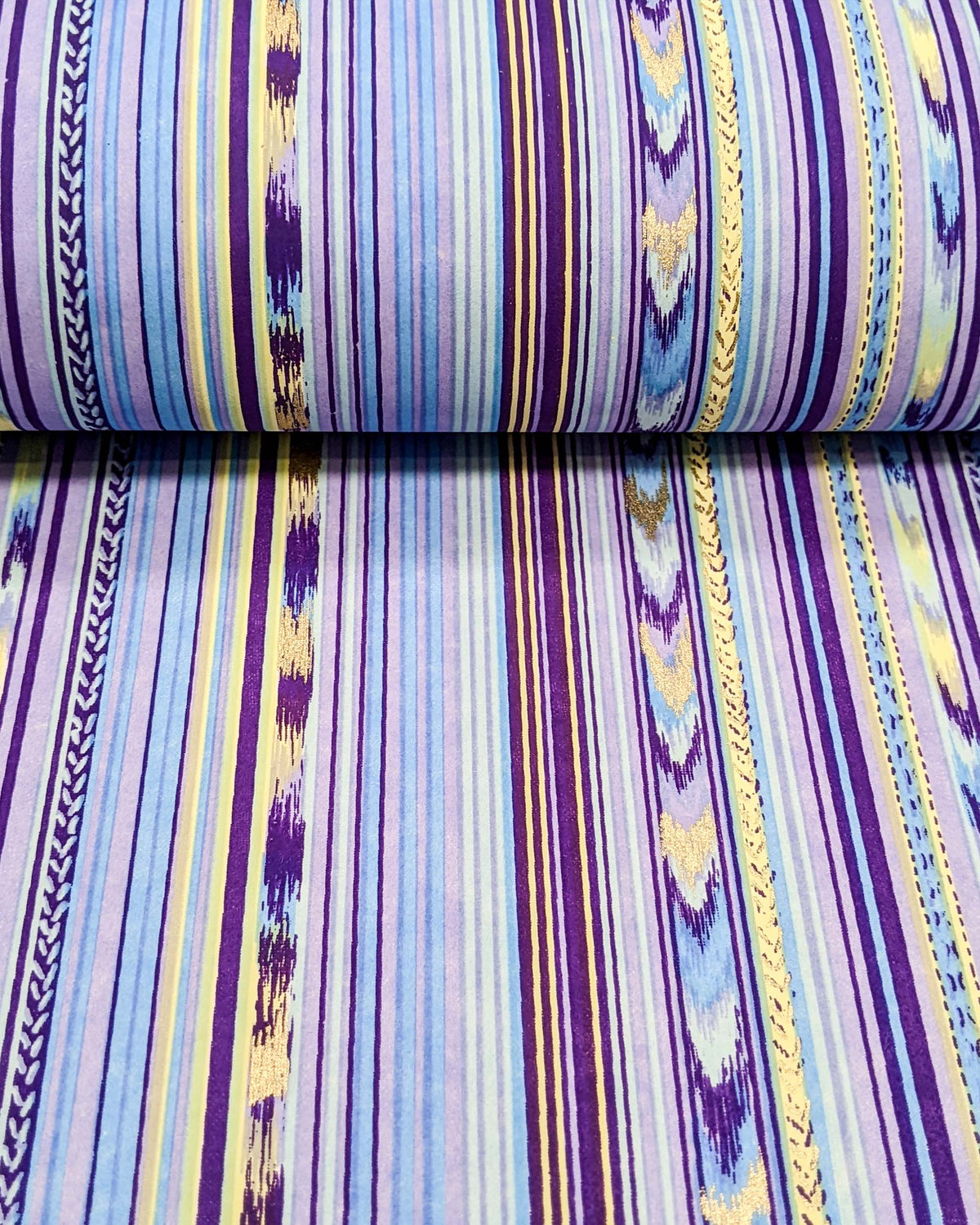 Japanese chiyogami paper featuring assorted solid and patterned vertical stripes in shades of purple, light blue, and yellow with metallic gold detail, shown rolled to highlight the design.