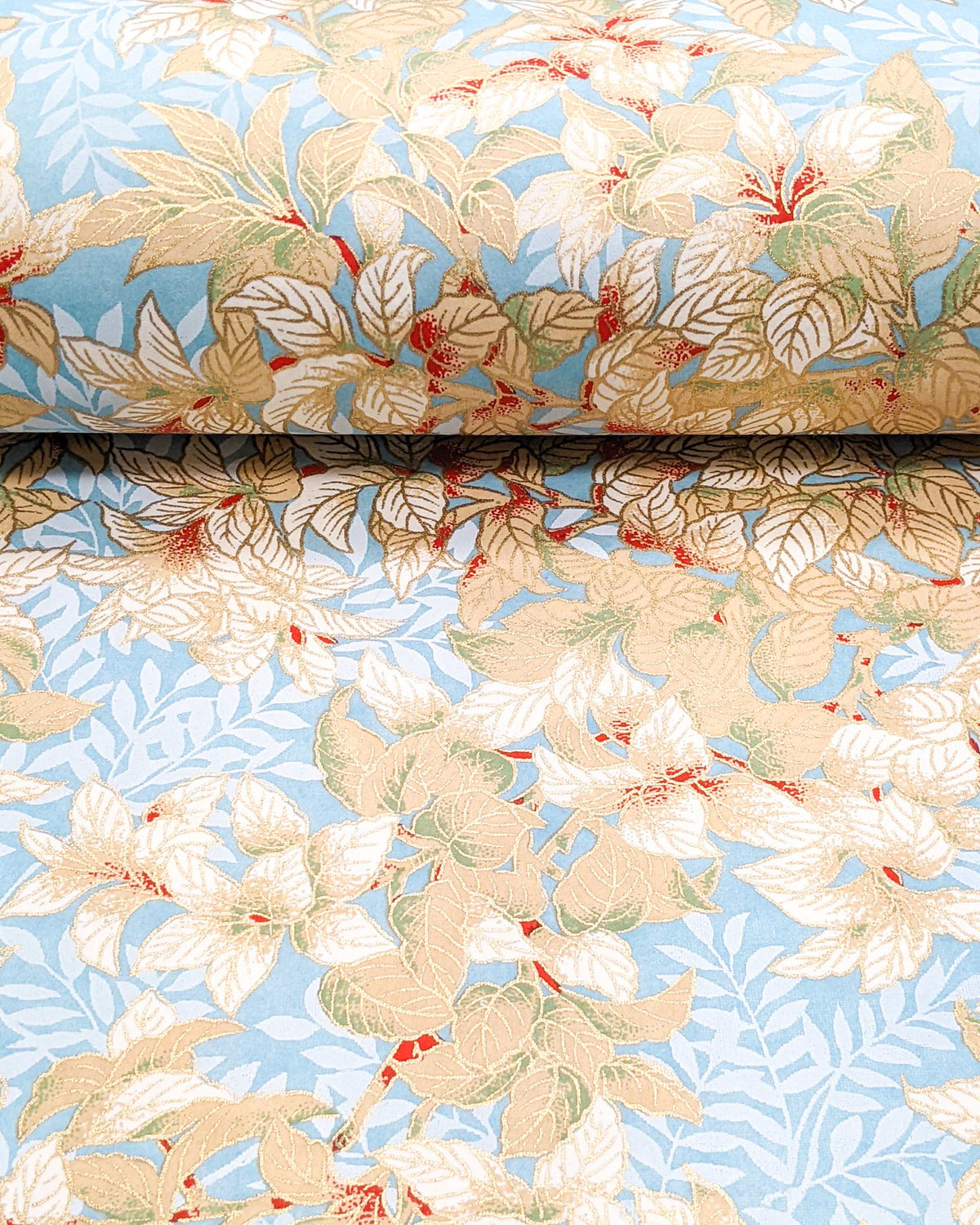 Japanese chiyogami paper featuring beige and white leaves with light blue leaves on a light blue background with metallic gold details, shown rolled to highlight the design.