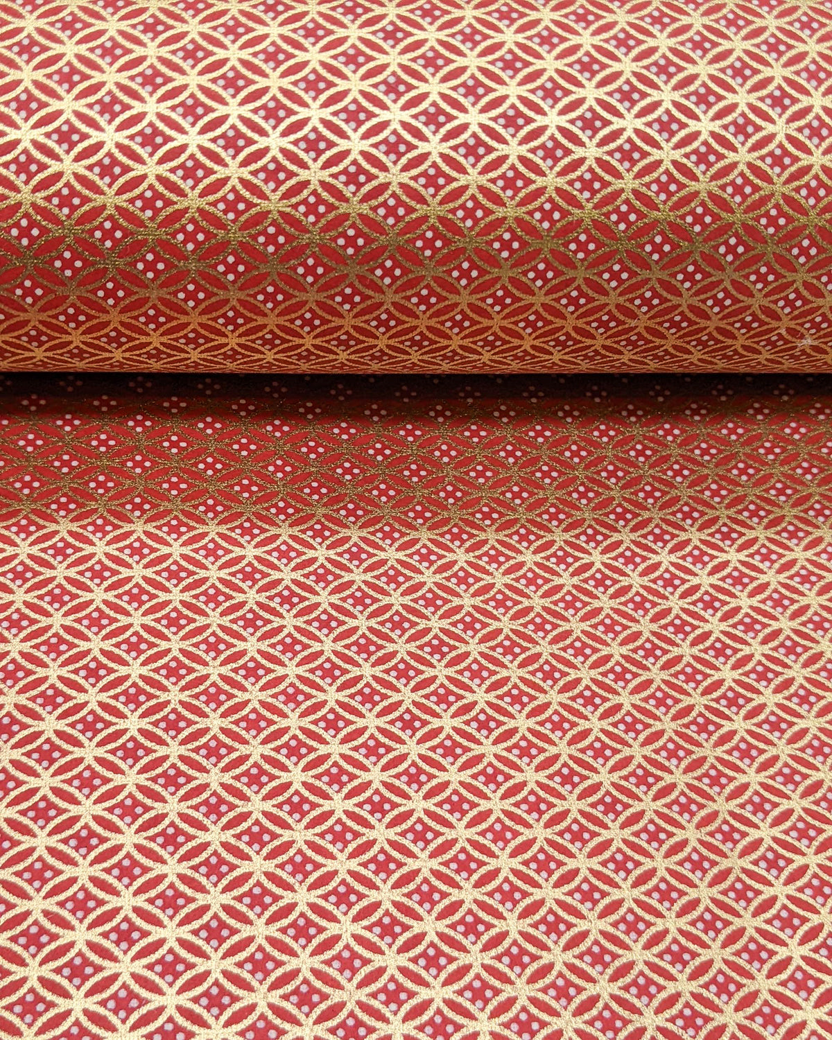 Japanese chiyogami paper featuring a traditional oval shippo pattern in metallic gold with four white dots in the center on a red background, shown rolled to highlight the design.