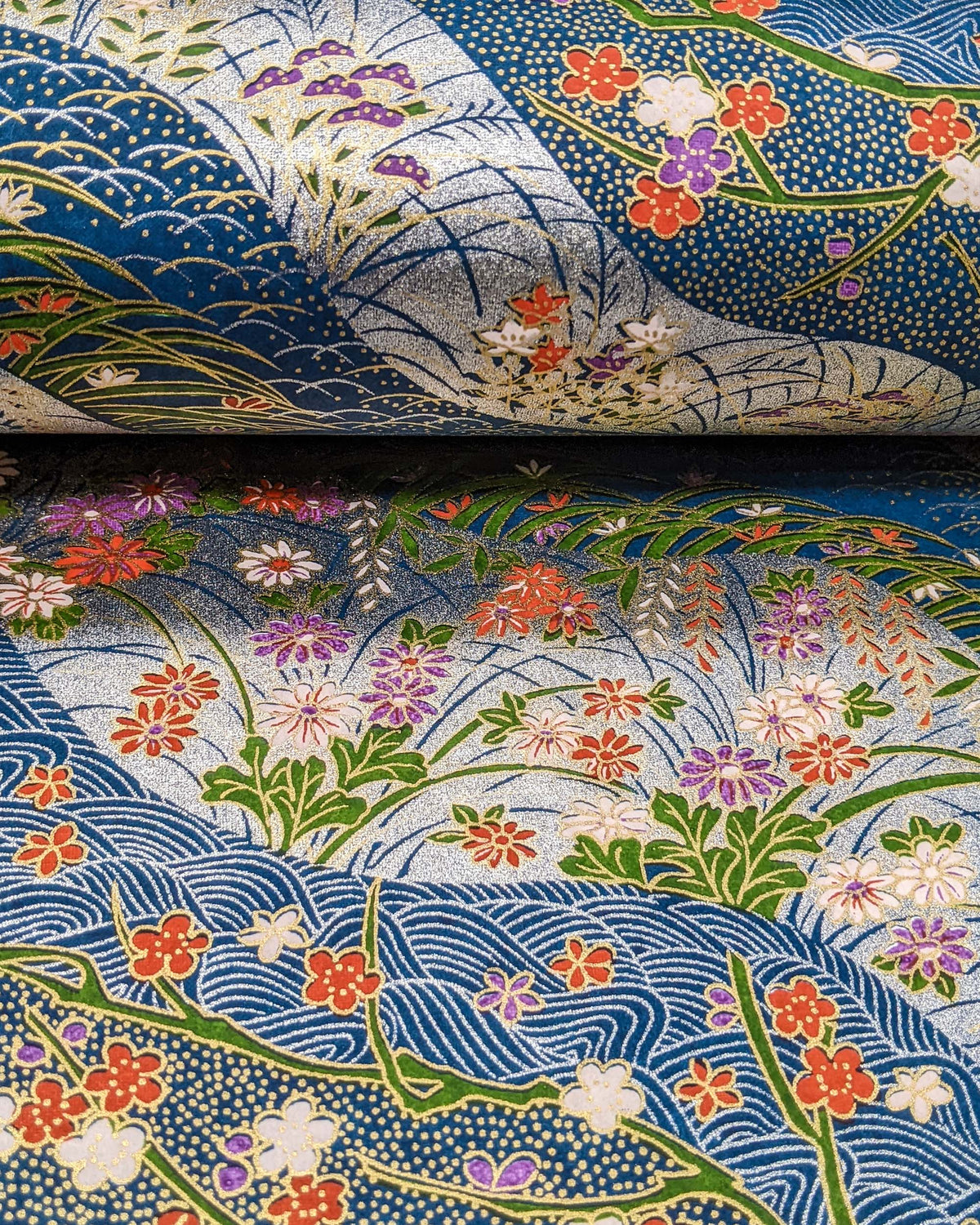 Japanese chiyogami paper featuringred, purple, pink, and white chrysanthemums, plum blossoms, wisteria, and other flowers arranged among traditional Japanese patterns on a blue and silver background with metallic gold details, shown rolled to highlight the design.