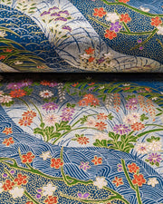 Japanese chiyogami paper featuringred, purple, pink, and white chrysanthemums, plum blossoms, wisteria, and other flowers arranged among traditional Japanese patterns on a blue and silver background with metallic gold details, shown rolled to highlight the design.